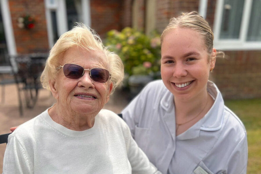 Older woman wearing sunglasses sits next to a young carer smiling outdoors, with a garden and brick building visible in the background.