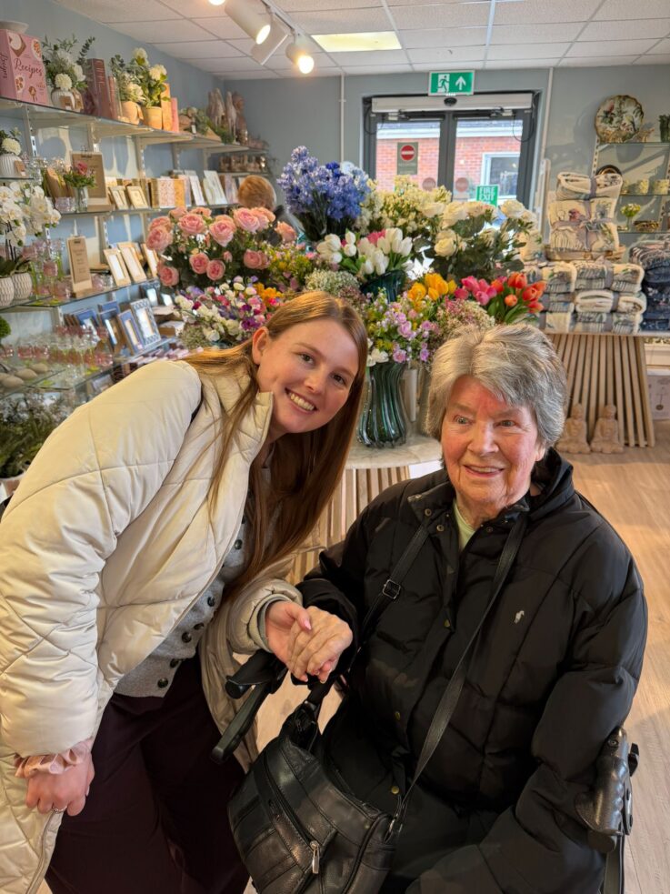 Two women smiling and holding hands inside a flower shop, with colourful floral arrangements and gift items displayed in the background.