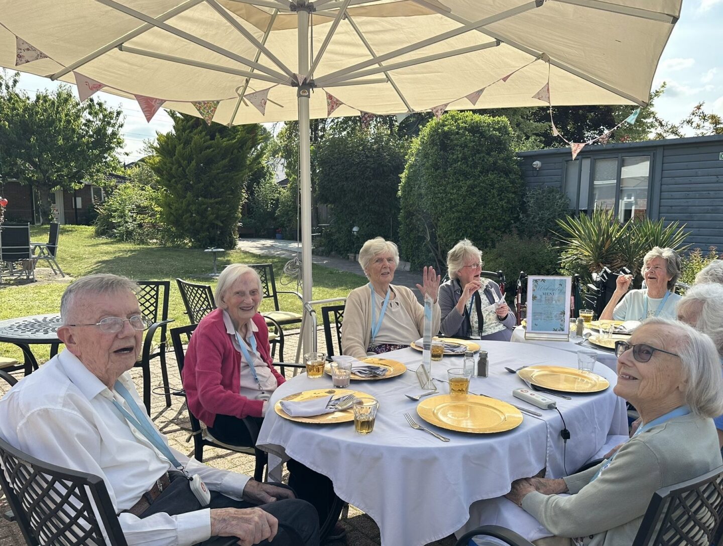 Six older adults sit around a table outdoors under a large parasol, with yellow plates and drinks, smiling and enjoying a meal together on a sunny day.