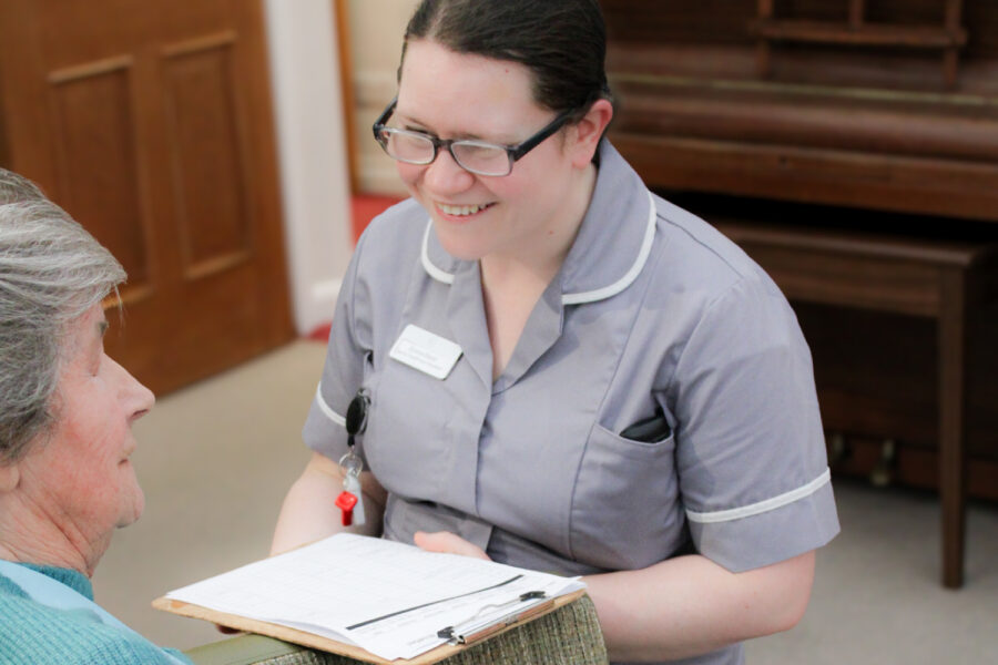 A nurse in uniform holds a clipboard and smiles whilst interacting with an elderly person seated in a chair.