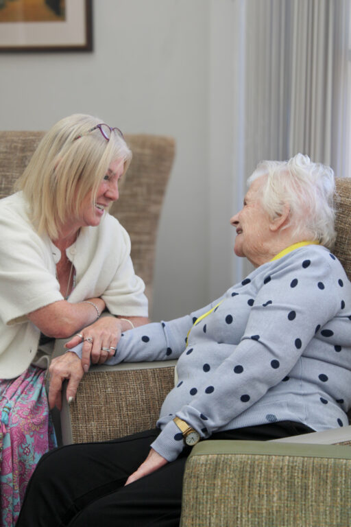 An older woman in a polka dot jumper sits and smiles whilst holding hands with another woman, who is leaning in and smiling back at her.