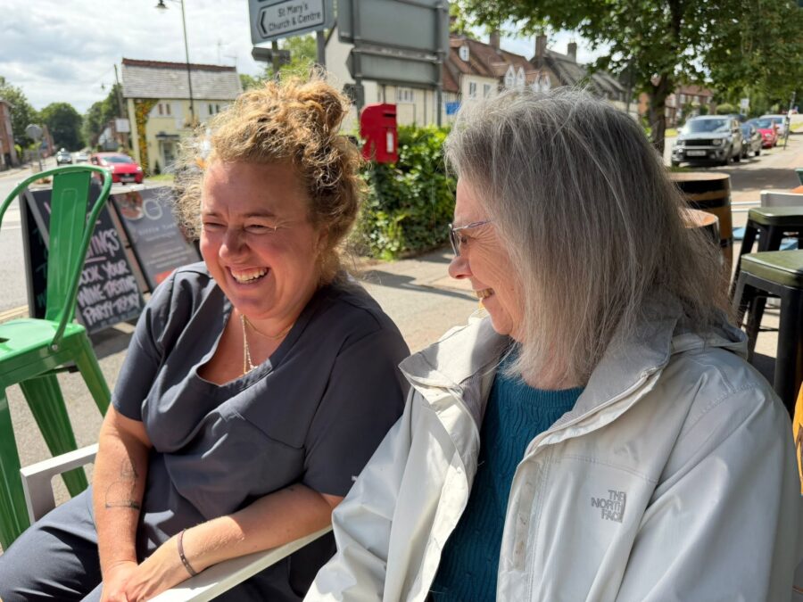 Two women sit outdoors at a table, smiling and talking, with sunlight shining on them; a street and shops are visible in the background.