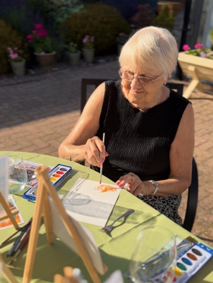 An older woman with glasses paints at a table outdoors, surrounded by watercolour supplies, canvases, and wine glasses on a sunny day.