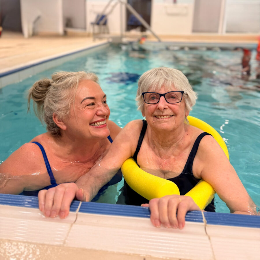 Two older women in swimsuits smile whilst holding onto the edge of an indoor swimming pool. One woman uses a yellow woggle for support.