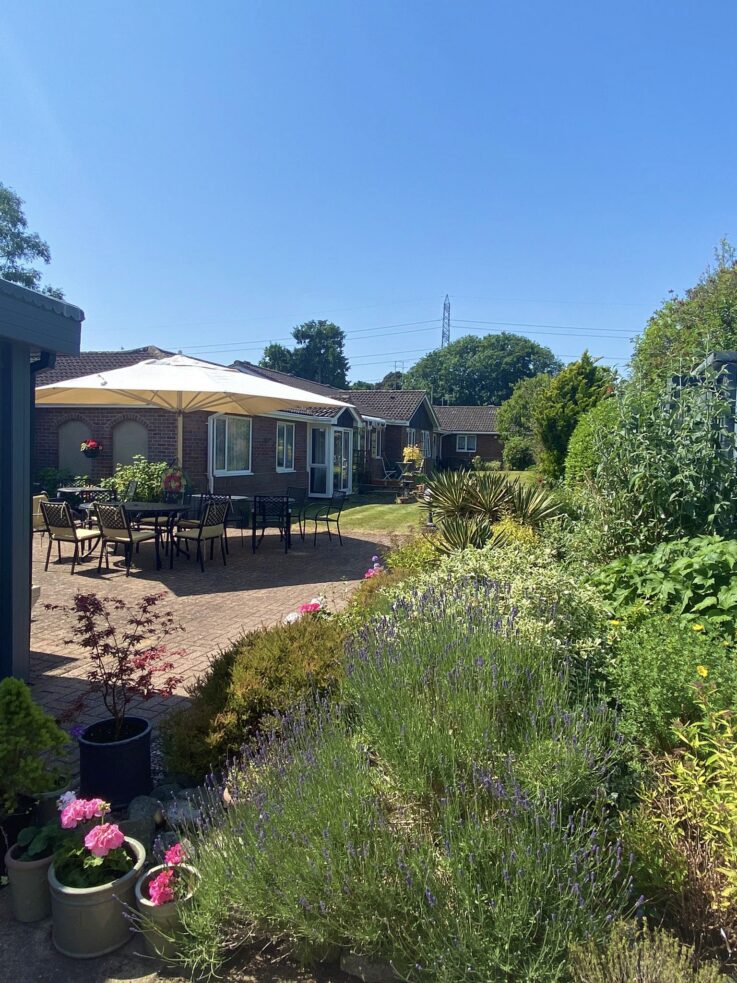 A sunny garden with flowering plants, patio tables and chairs under a large parasol, and houses in the background.