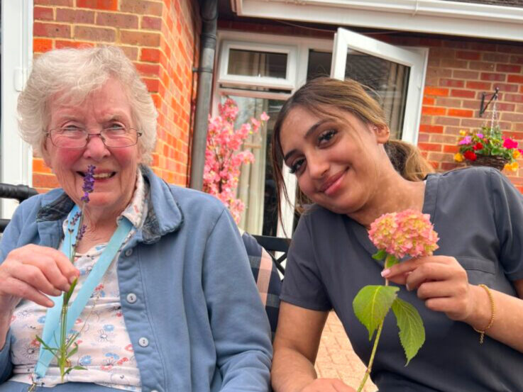 An older woman and a younger woman sit outdoors, each holding a flower and smiling. Brick building and potted plants are visible in the background.