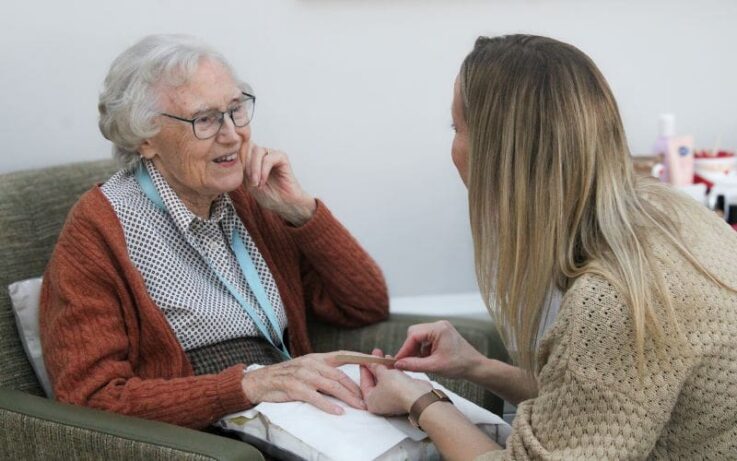 An elderly woman sits in a chair smiling whilst a younger woman gently holds and looks at her hand during a conversation.