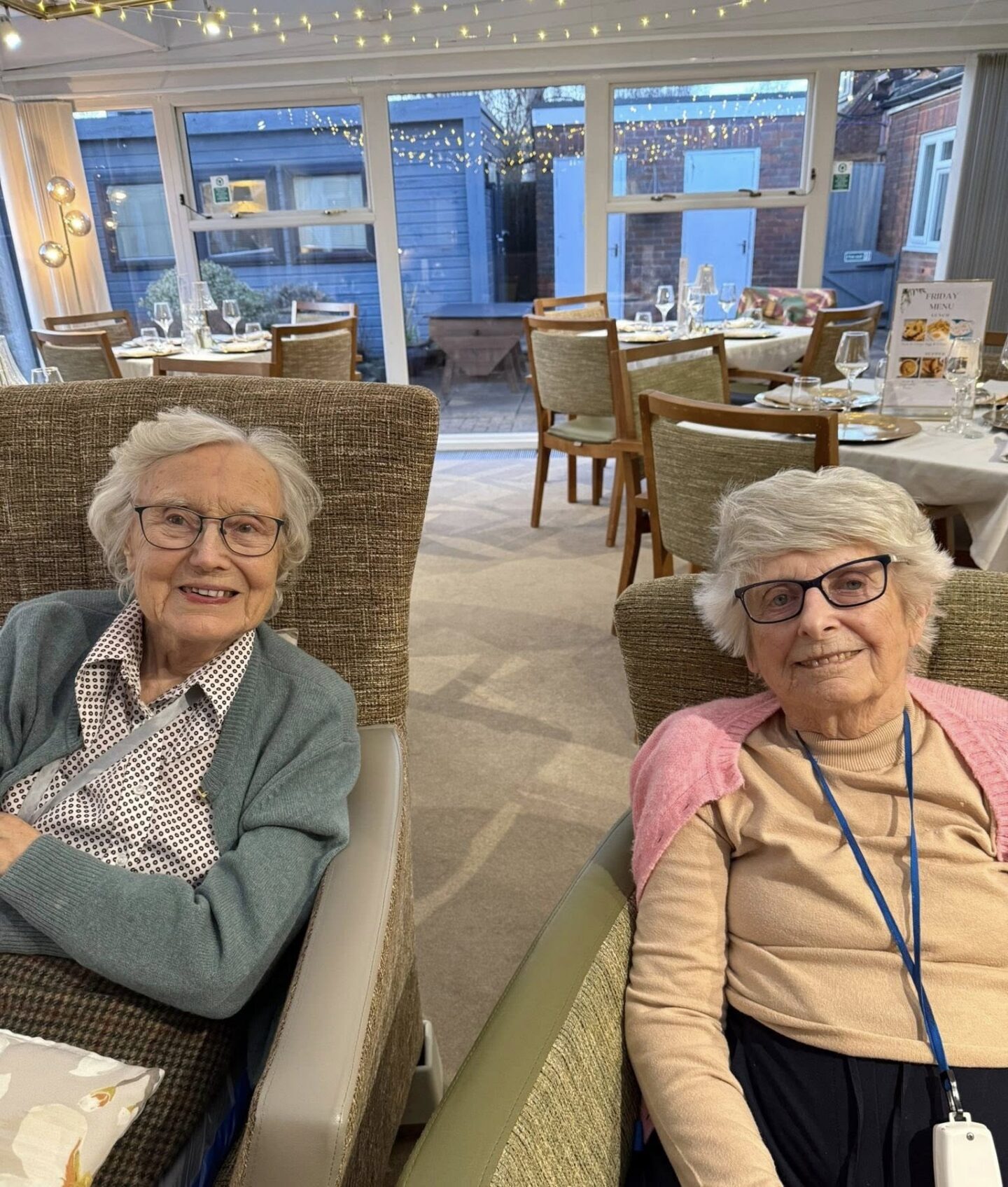 Two elderly women sit side by side in armchairs, smiling at the camera in a well-lit dining area with tables and chairs in the background.