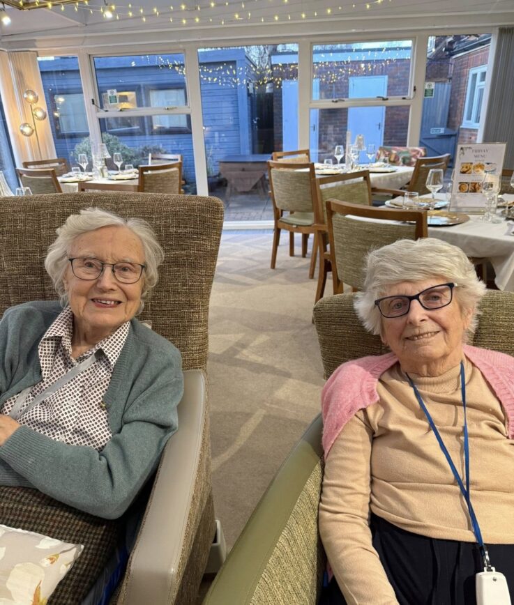 Two elderly women sit side by side in armchairs, smiling at the camera in a well-lit dining area with tables and chairs in the background.