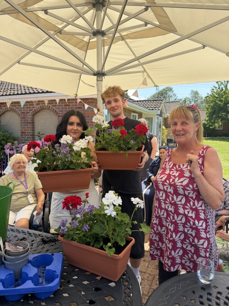 Three people under a garden umbrella hold pots of red and white flowers, with others seated in the background at an outdoor gathering.