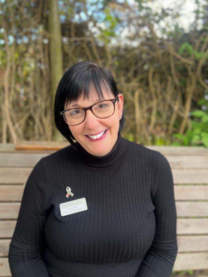 A person with short dark hair and glasses, wearing a black polo neck and name badge, smiles whilst sitting on a wooden bench outdoors.