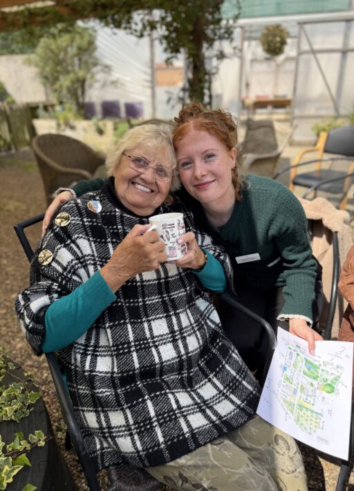 An older woman in a checked poncho holds a mug and smiles beside a younger woman holding papers with drawings, both seated outdoors.