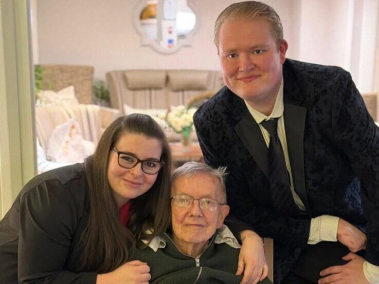 An elderly man sits in a chair, smiling, with a younger woman and a younger man standing close on either side, posing for a photo indoors.