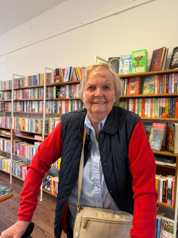 An older woman with short hair, wearing a red jumper and dark waistcoat, stands and smiles inside a bookshop filled with colourful books on shelves.