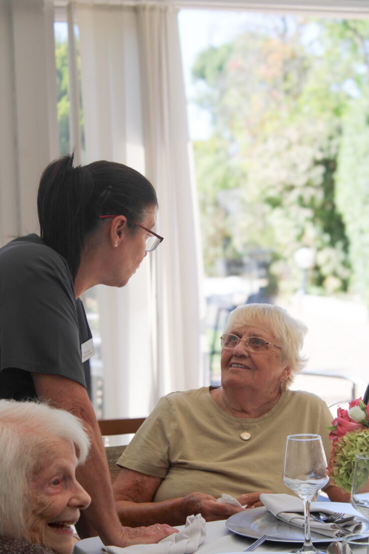A carer stands and speaks with an elderly woman seated at a dining table set with plates, glasses, and flowers, whilst another older woman smiles nearby.