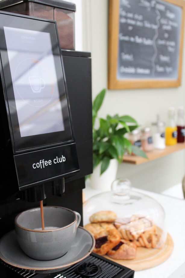 A coffee machine dispenses coffee into a grey cup, with pastries and a covered cake stand in the background on a counter.