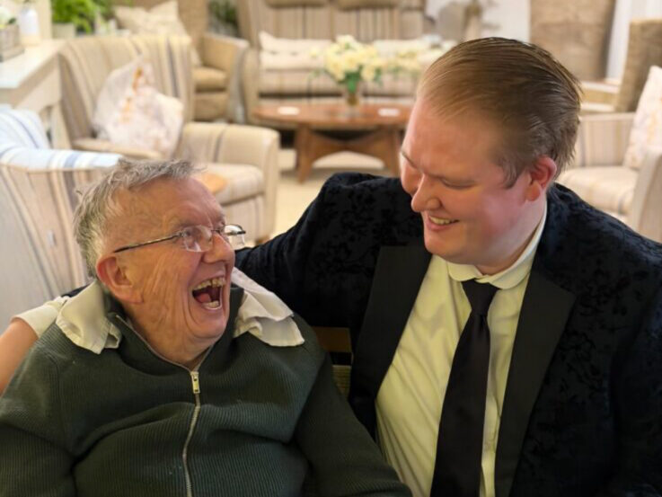 An elderly person and a younger adult in a suit sit together indoors, smiling and laughing, with arm round shoulders.