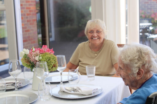 Two elderly women sit at a dining table set with plates, glasses, and a floral centrepiece, smiling in a bright room with large windows.