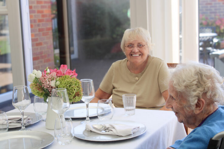 Two elderly women sit at a dining table set with plates, glasses, and a floral centrepiece, smiling in a bright room with large windows.