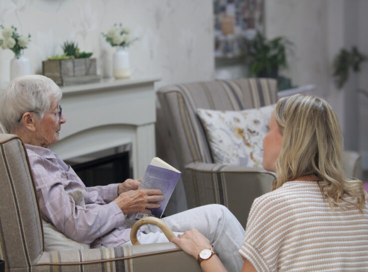 An elderly woman sits in an armchair reading a book whilst a younger woman sits nearby, listening attentively in a cosy living room.