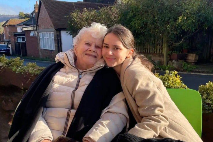 An elderly woman and a young woman sit closely together outdoors, smiling. Both are wearing light-coloured coats, and there are houses and greenery in the background.