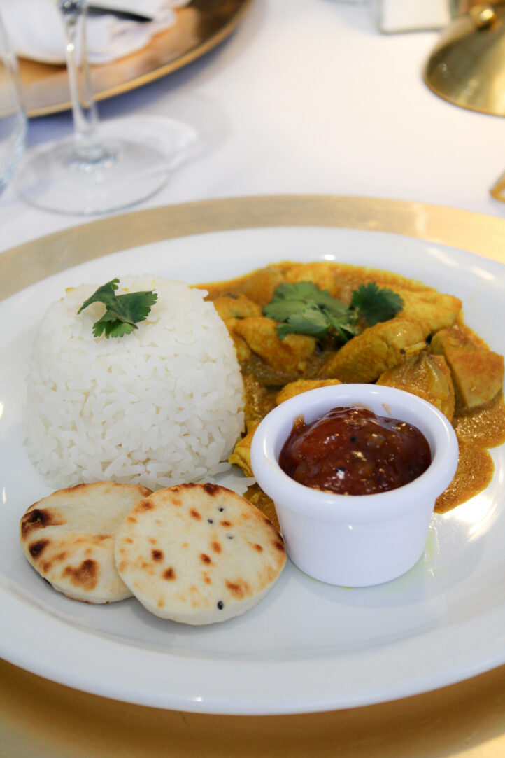 A plate with white rice, naan bread, a small pot of chutney, and a serving of curry garnished with coriander.