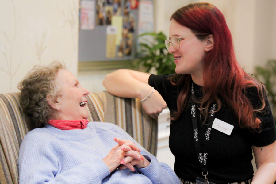 An elderly woman sits on a striped chair smiling at a young woman with glasses and a name badge, who is leaning on the chair; both appear to be engaged in conversation.