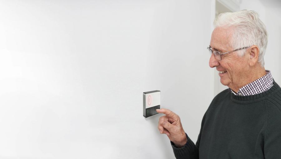 Older man with glasses pressing a button on a wall-mounted device, smiling whilst facing right in a bright, plain room.