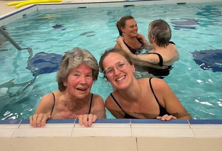 Two women smile at the edge of an indoor swimming pool while two pairs of women talk and exercise in the background. Pool equipment and a wheelchair are visible around the room.