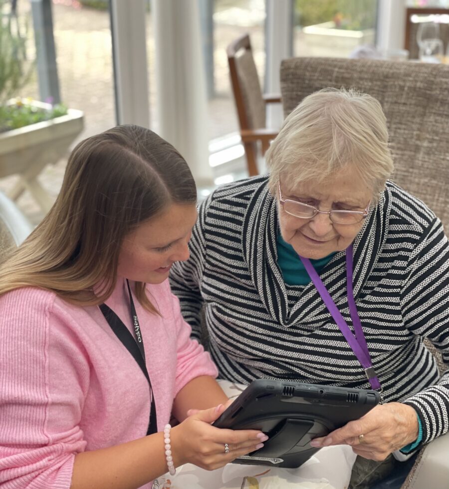 A younger woman helps an older woman use a tablet device while sitting together indoors.
