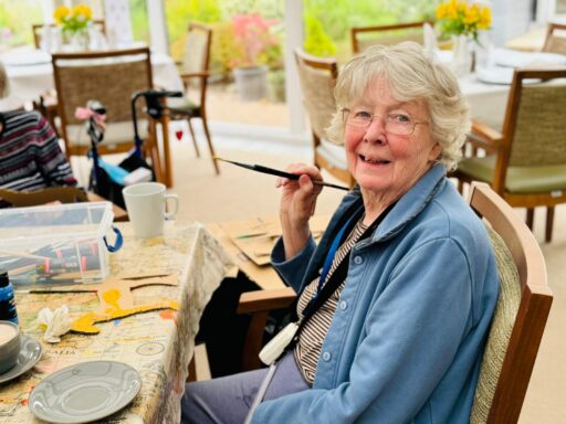 An elderly woman sits at a table holding a paintbrush, surrounded by art supplies, smiling at the camera in a bright, sunlit room.