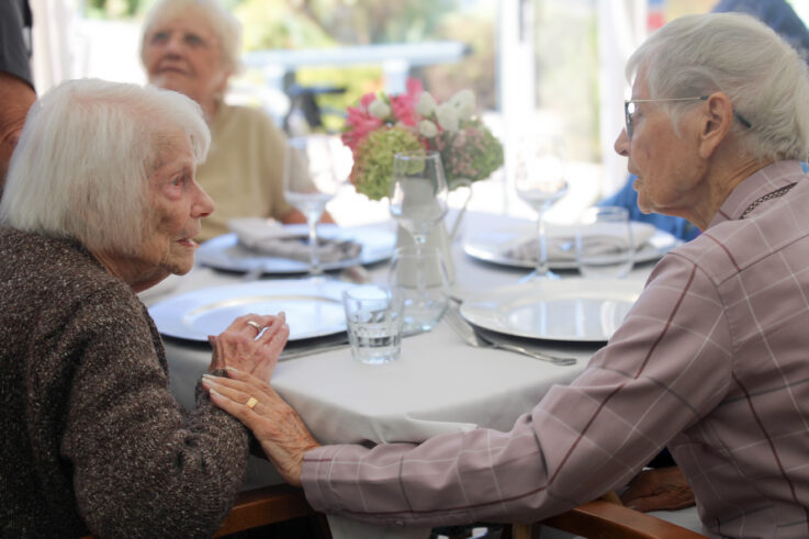 Two elderly women sit at a round table set for a meal, holding hands and talking, with two others seated in the background.