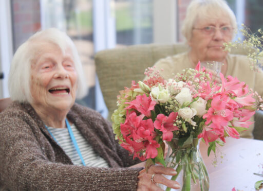 Two elderly women sit indoors at a table with a vase of pink and white flowers. One woman is smiling, whilst the other looks forward.