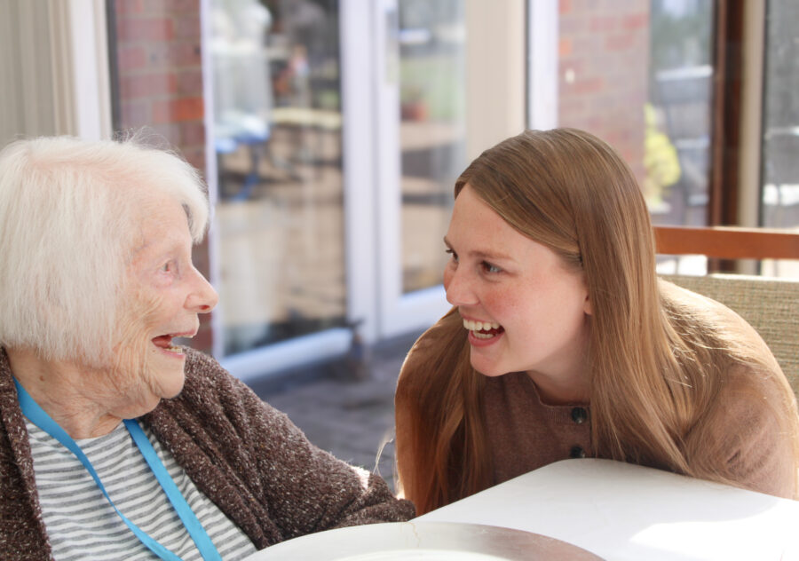 An elderly woman and a younger woman sit together at a table, smiling and looking at each other in a well-lit room with large windows in the background.