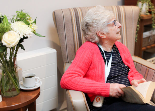 An elderly woman with grey hair and glasses sits in an armchair, holding an open book. A vase of flowers and a cup of coffee are on a nearby table.