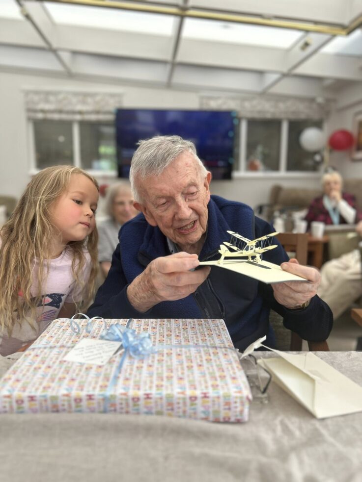 An elderly man examines a model aeroplane while a young girl watches. A wrapped present and card are on the table, with other people and decorations in the background.
