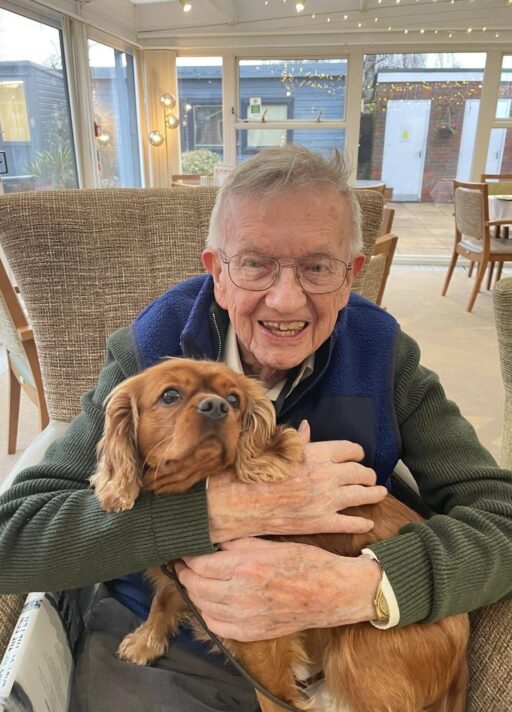 Elderly man sitting in an armchair indoors, smiling and holding a brown dog on his lap.