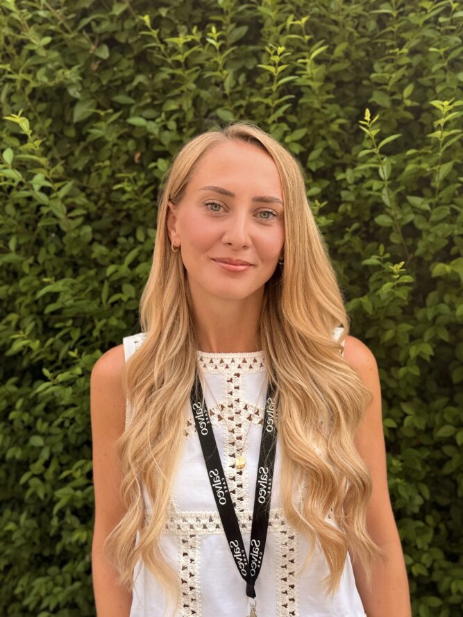 A woman with long blonde hair wearing a white sleeveless top and a black lanyard stands in front of green leafy bushes.