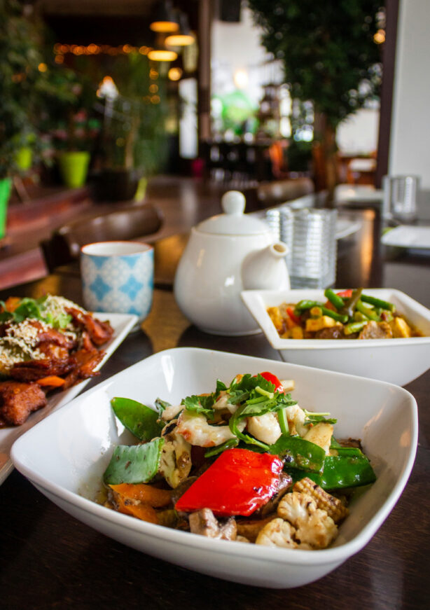 A white bowl of stir-fried vegetables and tofu on a table, with a teapot, cup, and additional dishes in the background at a restaurant.