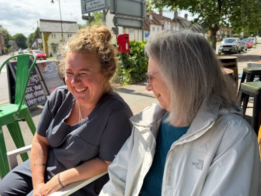 Two women sit outdoors at a café table, smiling and talking on a sunny day. There are chairs, street signs, and cars visible in the background.
