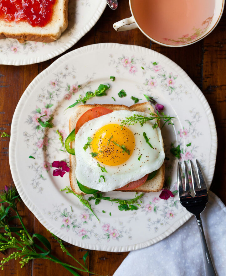 A plate with toast topped with tomato slices, greens, and a fried egg, next to a fork, with tea and jam on toast nearby.