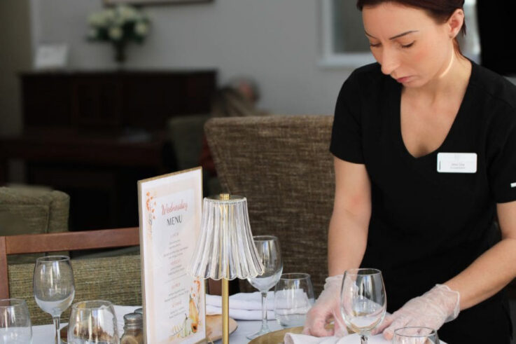 A woman in black uniform and gloves lays a dining table with glasses, next to a menu and lamp, in a well-lit indoor setting.