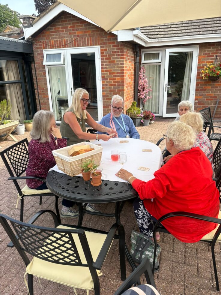 Five older adults sit around a round patio table outdoors, engaged in conversation, with drinks and a basket on the table.