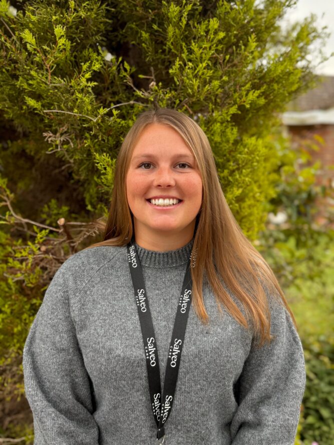 A woman with long hair and a grey jumper, wearing a lanyard that says Salford, stands outdoors in front of a green bush.