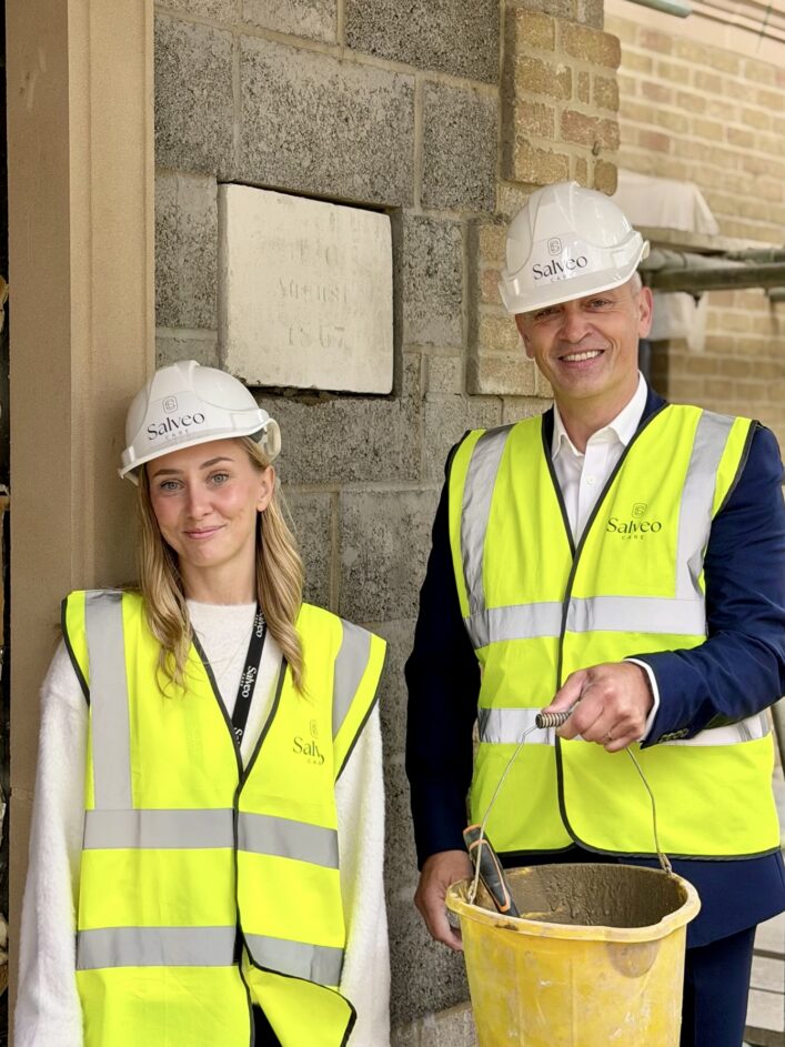 A woman and a man wearing white Salveo Care hard hats and high-visibility vests stand before a stone wall, with the man holding a bucket of mortar.