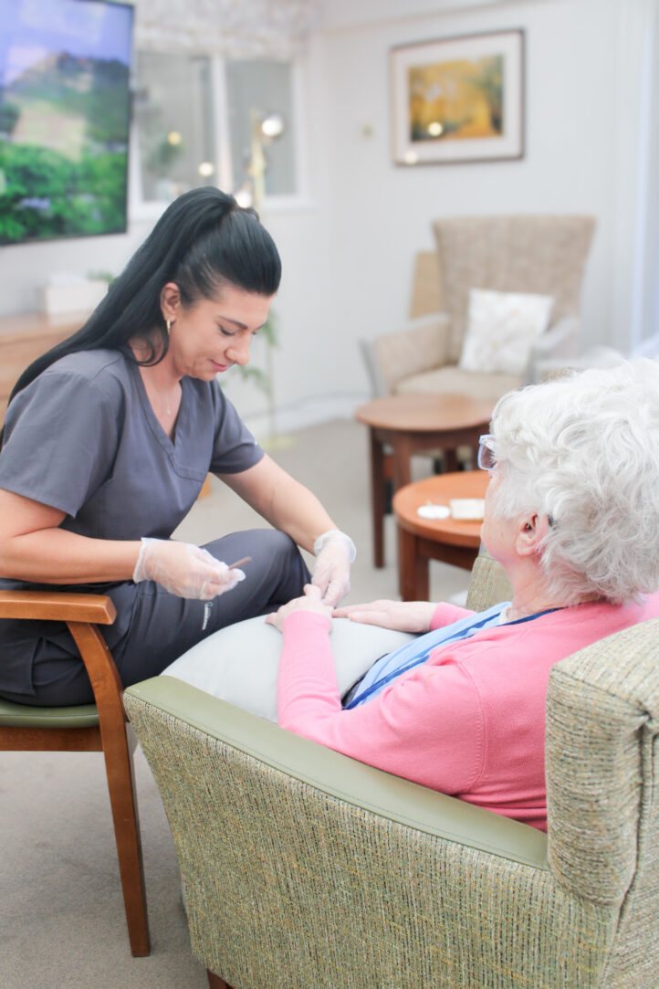 A carer in scrubs and gloves assists an elderly woman seated in a chair inside a well-lit lounge.