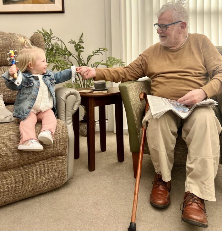 An elderly man sitting with a newspaper and walking stick reaches out to hold hands with a smiling toddler sitting on a nearby chair.