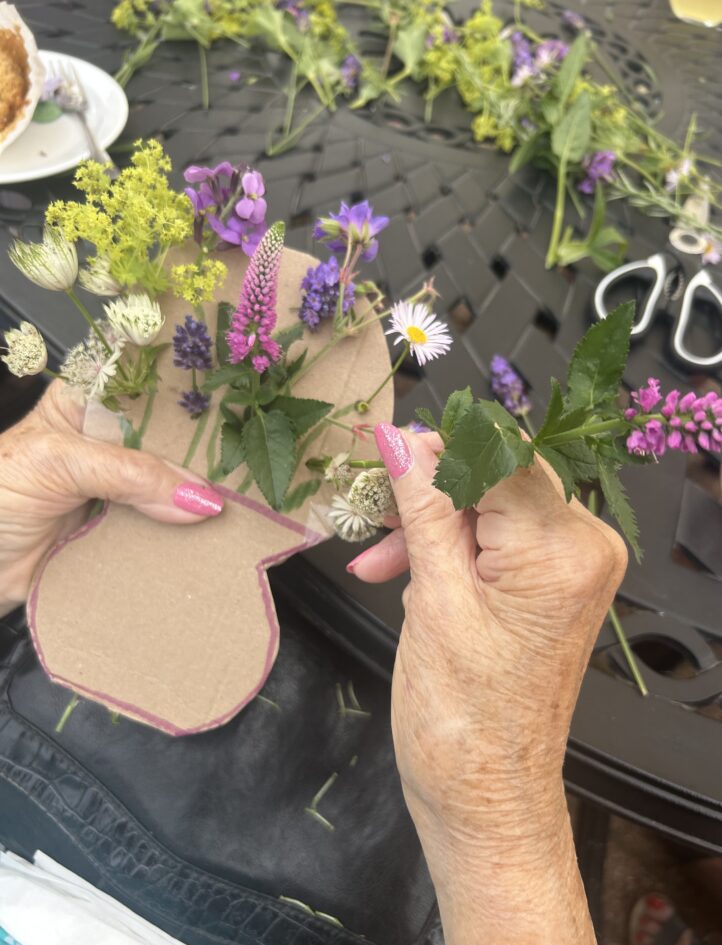 Hands arranging assorted wildflowers on a cardboard vase shape atop a black outdoor table, with scissors and more flowers in the background.