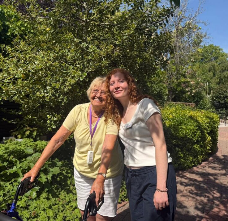 An older woman with a Zimmer frame and a younger woman stand close together, smiling, on a sunny path surrounded by greenery.