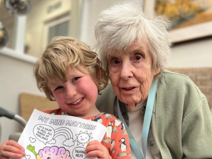 A smiling child holds a My Mind Matters colouring sheet next to an elderly woman with grey hair, both sitting indoors.